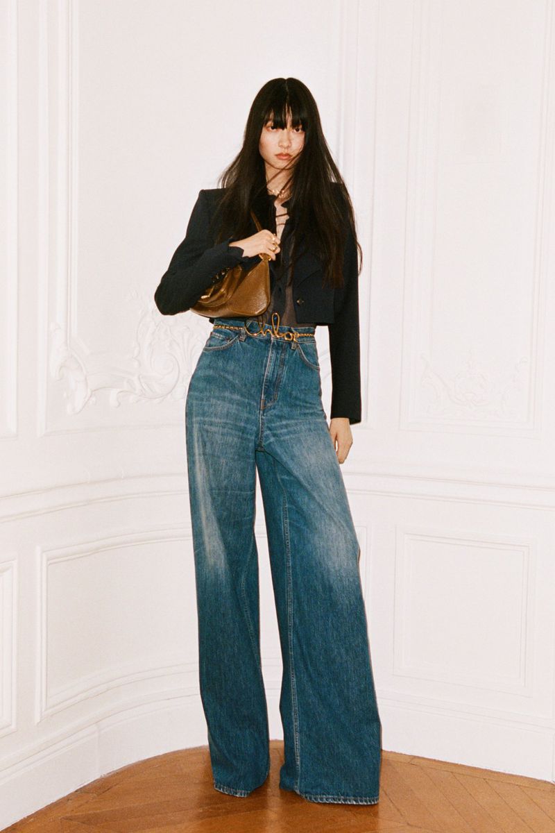 Full-length shot of a fashionable young Asian woman in flared jeans, a cropped jacket, and a shiny handbag against a neutral background.
