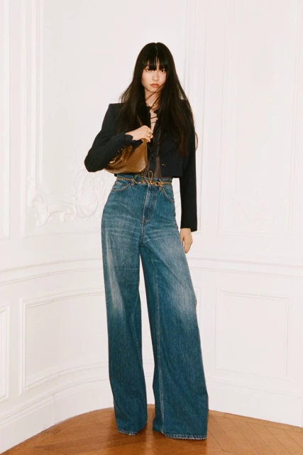 Full-length shot of a fashionable young Asian woman in flared jeans, a cropped jacket, and a shiny handbag against a neutral background.