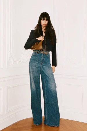 Full-length shot of a fashionable young Asian woman in flared jeans, a cropped jacket, and a shiny handbag against a neutral background.