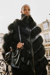 Model in a black textured shearling jacket holding a black leather handbag against an exterior balcony backdrop.