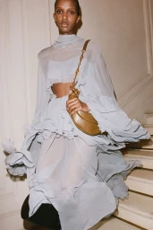 Model in a sheer pale blue ruffled dress holding a metallic gold shoulder bag standing on stairs