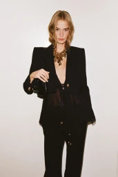 Model standing against a plain backdrop wearing a black cropped jacket with defined shoulders, sheer top, and chunky gold necklace.