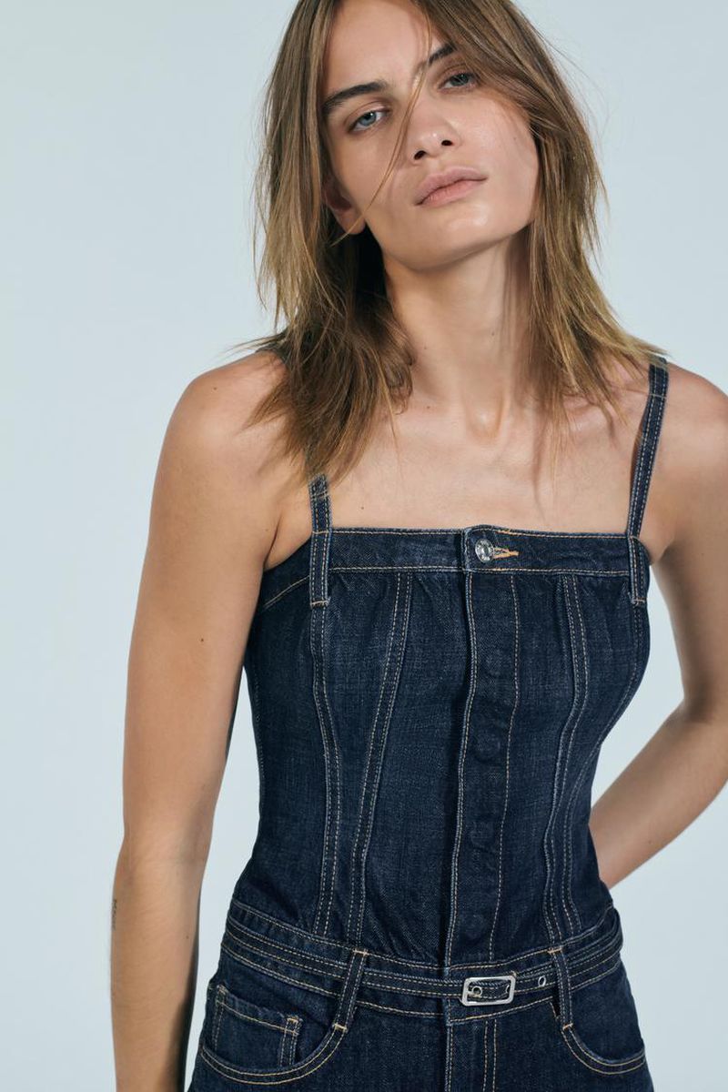 A woman poses in a denim one-piece outfit in a studio setting. The denim jumpsuit is buttoned and belted, complemented by her neutral expression.