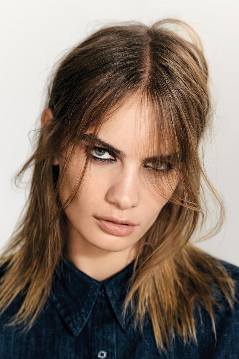 Close-up studio portrait of a young adult female with dark eyeliner, wearing a denim collared shirt and looking directly at the camera.