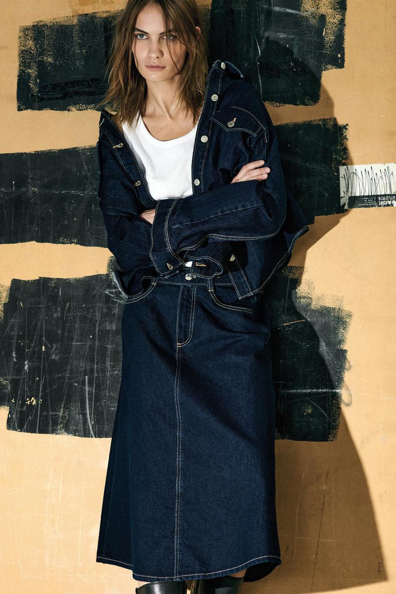 A woman poses against an abstract background, wearing a denim jacket and skirt, accessorized with a white t-shirt and dark boots.