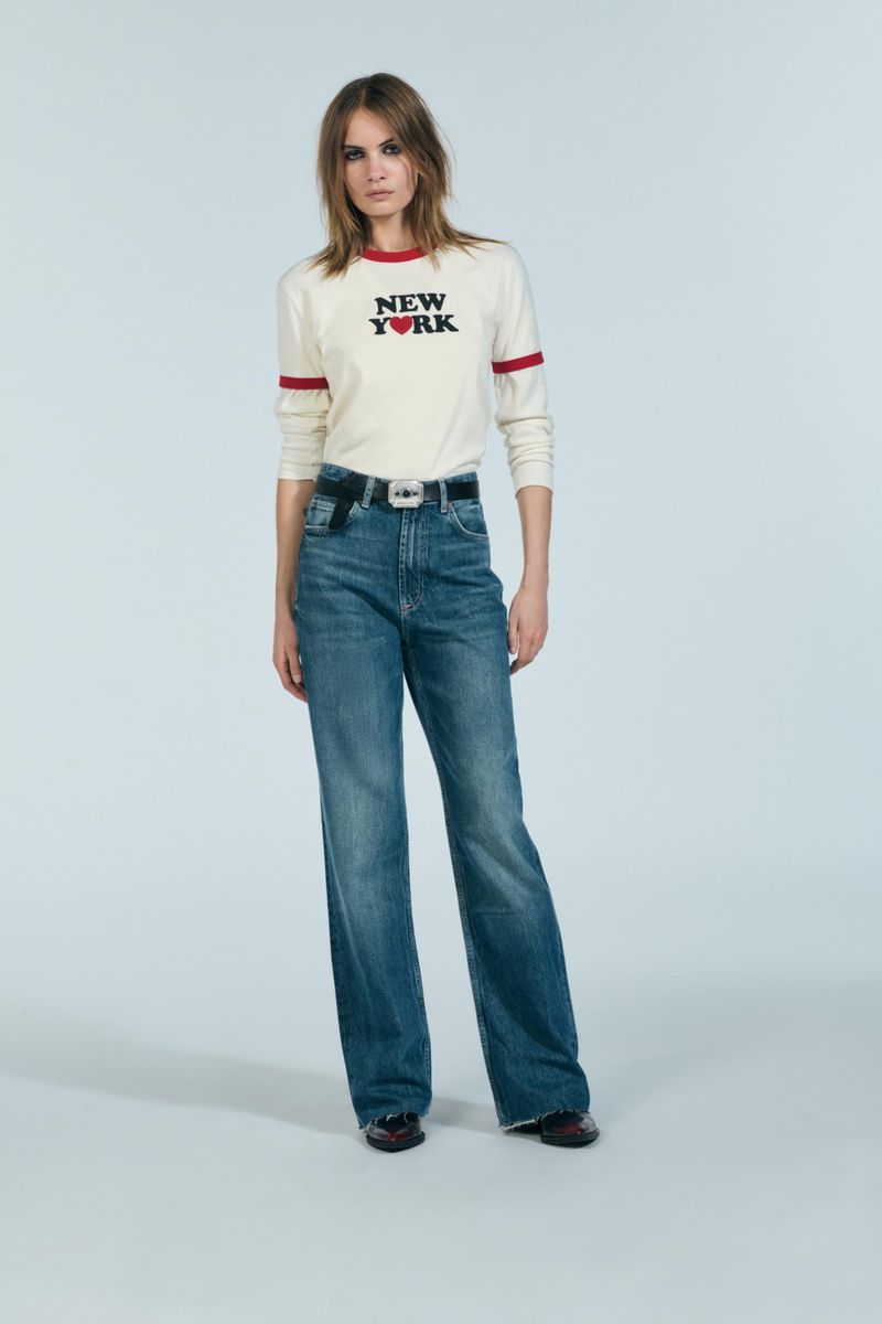 A woman in jeans and a t-shirt stands in a full shot against a light background, posing in a casual studio setting.