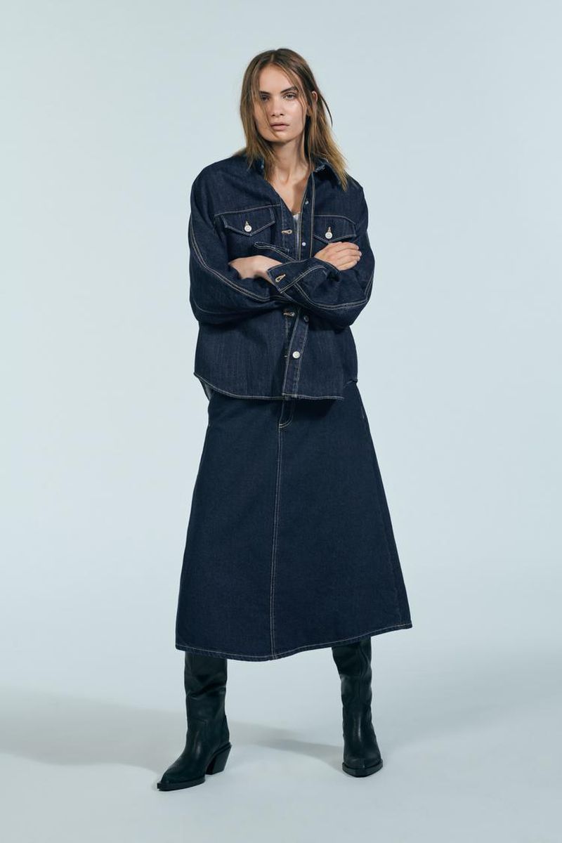 Model wearing dark denim shirt and long denim skirt with black boots against a plain light grey studio background.
