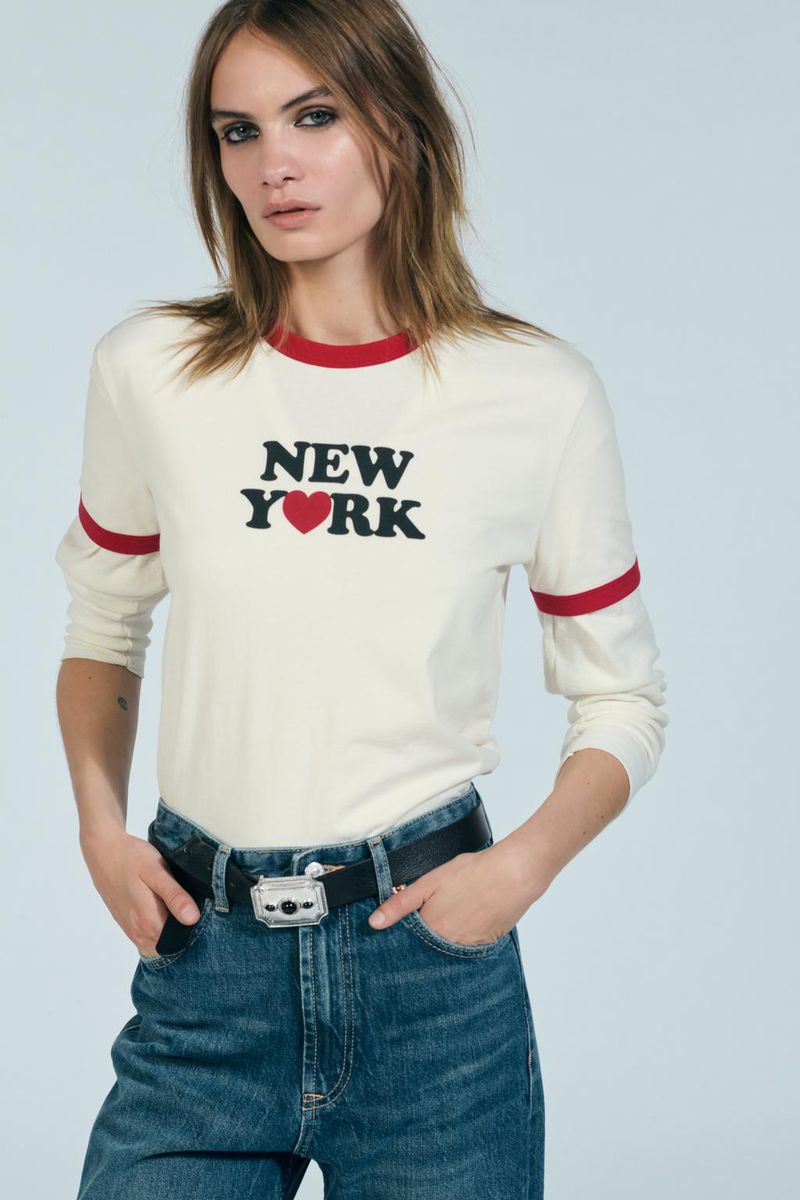 A woman in a long-sleeved shirt with 'New York' printed on it poses against a white background, hands in her jean pockets.
