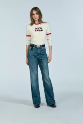 Model wearing a white New York long-sleeve ringer top and blue denim jeans against a plain grey background