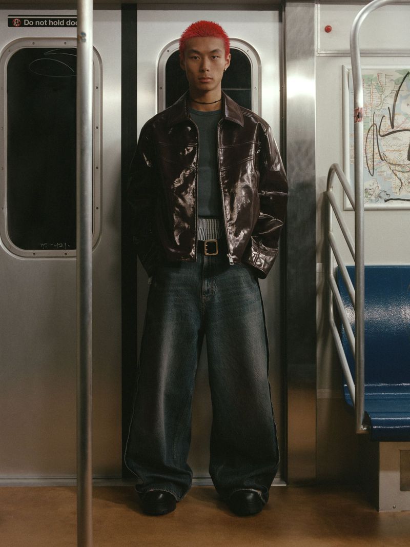 Young Asian man with bright red hair stands in wide-leg jeans and brown jacket in a subway car, looking directly at the camera.