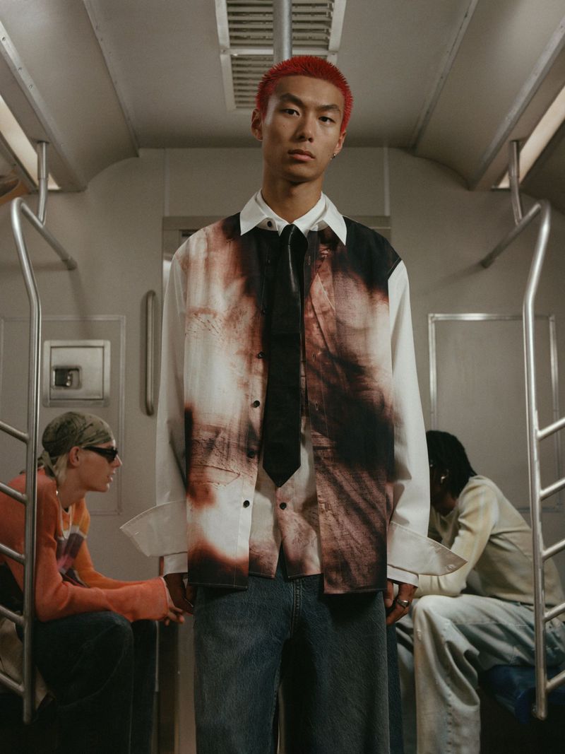Fashion shot in a subway with an adult male wearing an abstract print shirt and tie, alongside two other blurred people.