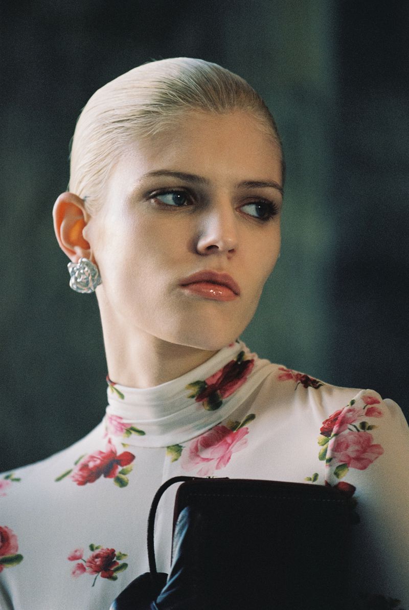Close-up studio portrait of a young adult female fashion model with slicked-back blonde hair, wearing a floral-print top and black gloves, looking away.