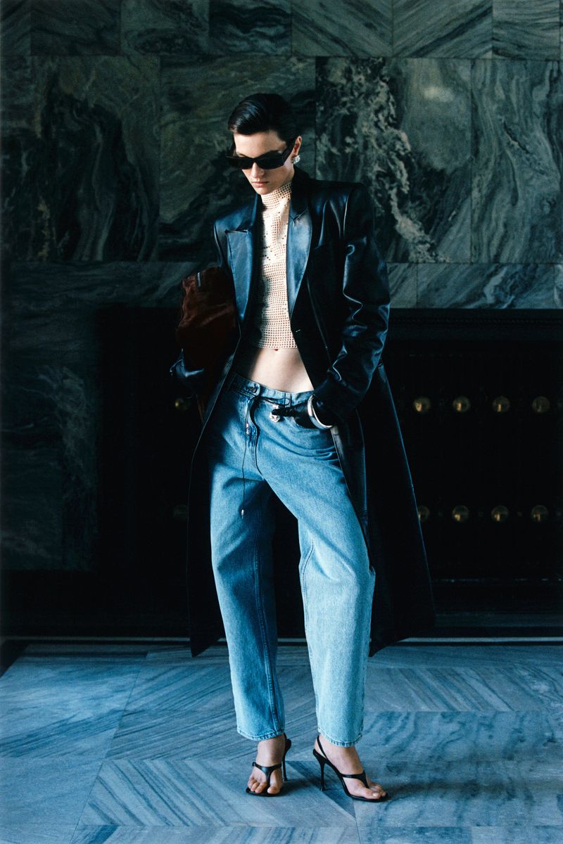 A fashionable adult woman wearing a long black coat and jeans poses in an indoor setting with a marbled wall in the background.