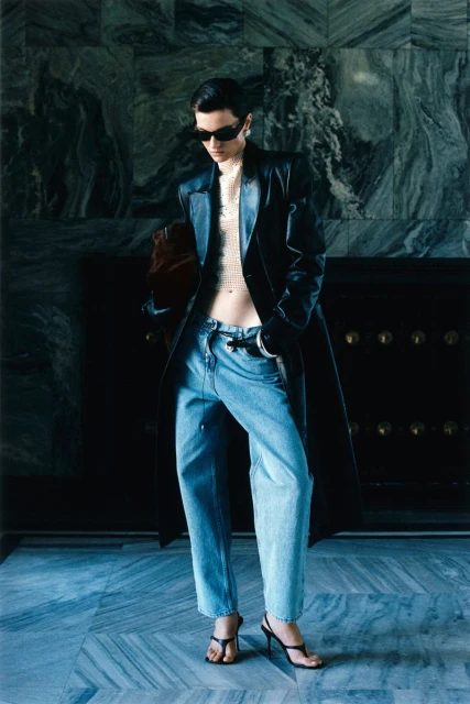 A fashionable adult woman wearing a long black coat and jeans poses in an indoor setting with a marbled wall in the background.
