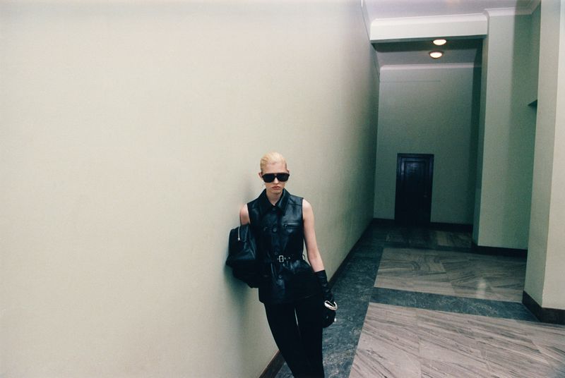 Stylish woman with blonde hair poses in a black leather vest, slacks, sunglasses, and gloves indoors, standing against a wall.