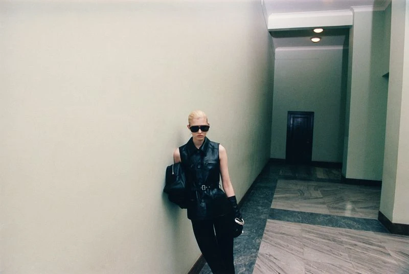 Stylish woman with blonde hair poses in a black leather vest, slacks, sunglasses, and gloves indoors, standing against a wall.