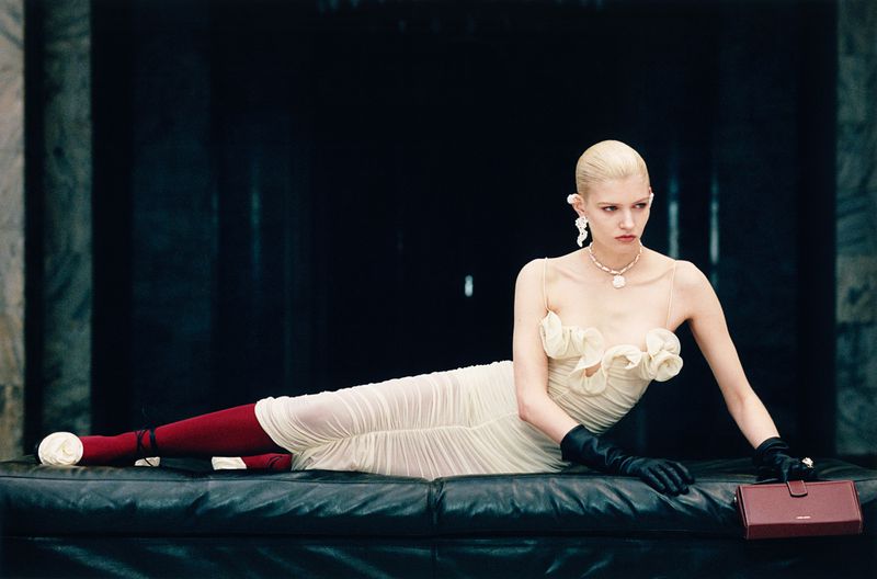 A poised fashion model lounges on a black leather bench wearing an elegant cream dress with floral details, jewelry, gloves, and red tights.