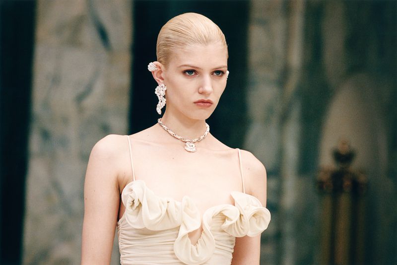 Close-up of a fair-skinned woman with slicked-back blonde hair wearing a ruffled cream dress and ornate jewelry in a muted indoor setting.