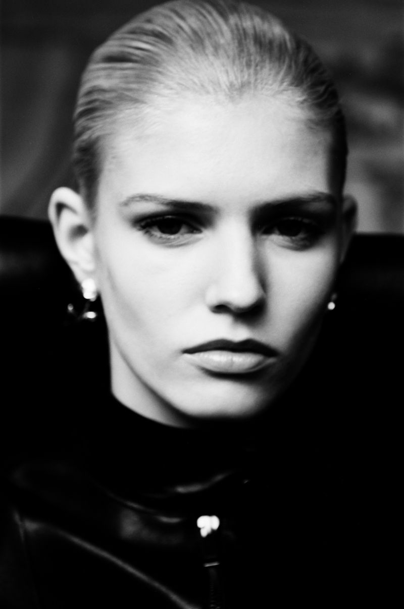 Close-up portrait of an adult female with slicked-back blonde hair wearing a black top in a studio setting with neutral facial expression.