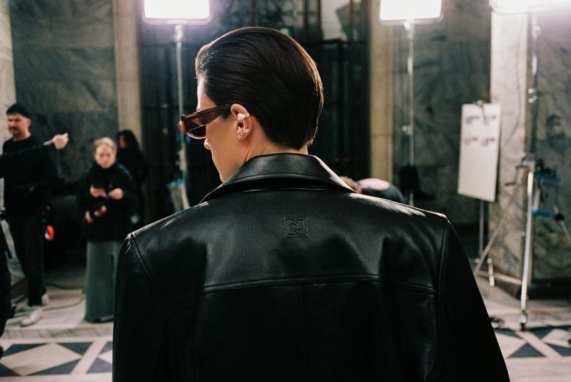 Woman in black leather jacket at a photoshoot, captured from behind, with crew and lighting equipment visible in the background.