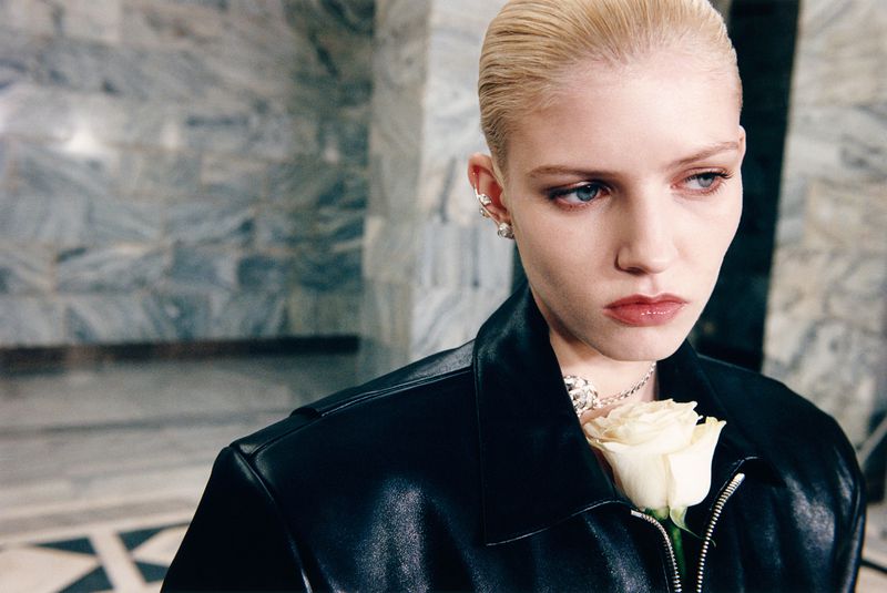 Close-up shot of a fair-skinned woman with short blonde hair wearing a black leather jacket and silver jewelry holding a white rose.