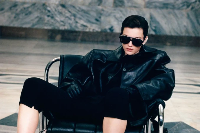 Model Camille Chifflot reclining in a chrome armchair wearing an oversized black leather jacket and dark sunglasses.
