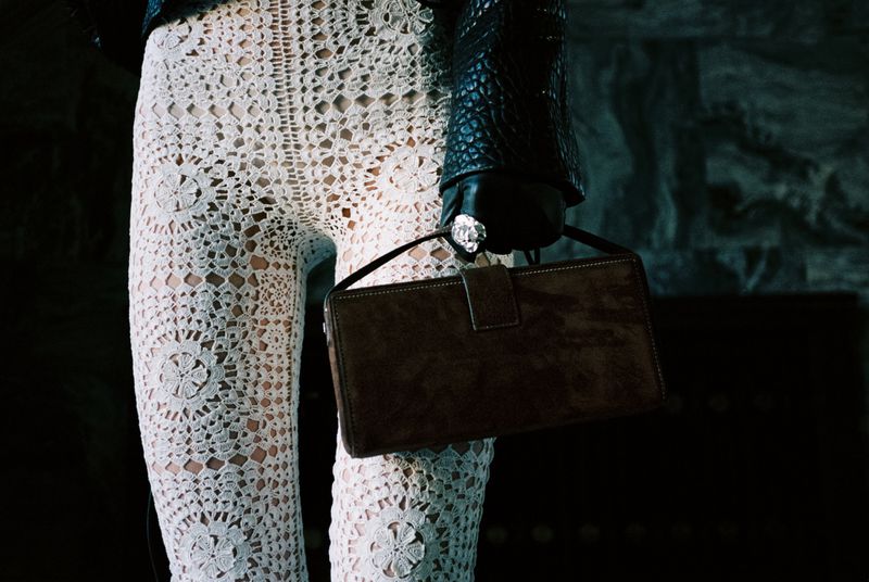 Close-up of person wearing white crochet pants, black leather jacket, gloves with ring, and holding brown handbag in dim interior.
