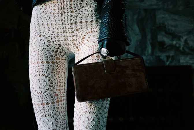 Close-up of person wearing white crochet pants, black leather jacket, gloves with ring, and holding brown handbag in dim interior.