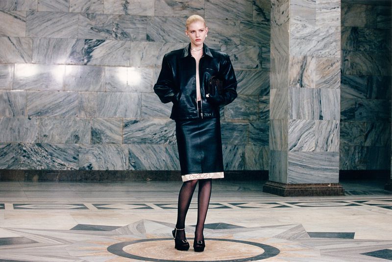 An adult female fashion model poses in a black leather jacket and pencil skirt in an indoor environment with a marble background.
