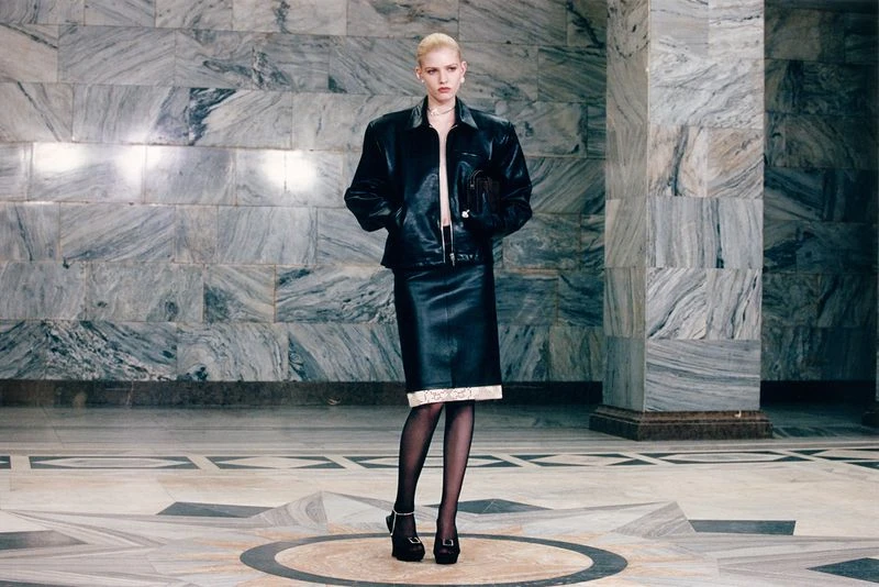 An adult female fashion model poses in a black leather jacket and pencil skirt in an indoor environment with a marble background.