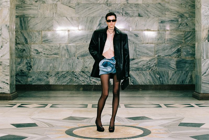 Full-length studio shot of a female model in a black jacket, light crop top, denim skirt, black stockings, and platform heels striking a pose.