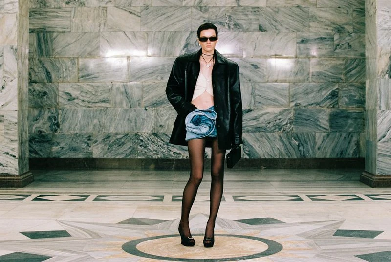 Full-length studio shot of a female model in a black jacket, light crop top, denim skirt, black stockings, and platform heels striking a pose.