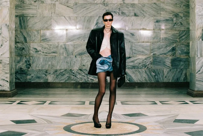 Full-length studio shot of a female model in a black jacket, light crop top, denim skirt, black stockings, and platform heels striking a pose.