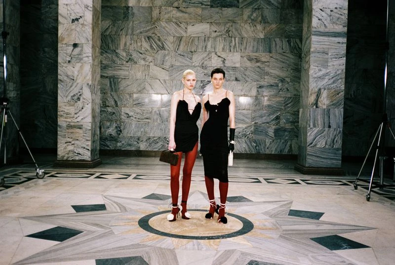 Two women in black dresses pose in an elaborate marble hall. One carries a handbag, and the other a bottle, with a spotlight visible.