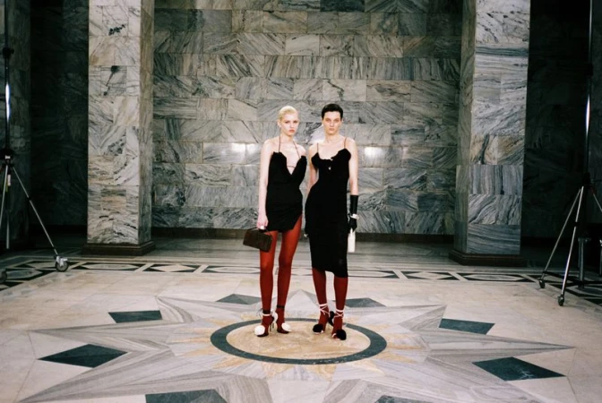 Two women in black dresses pose in an elaborate marble hall. One carries a handbag, and the other a bottle, with a spotlight visible.