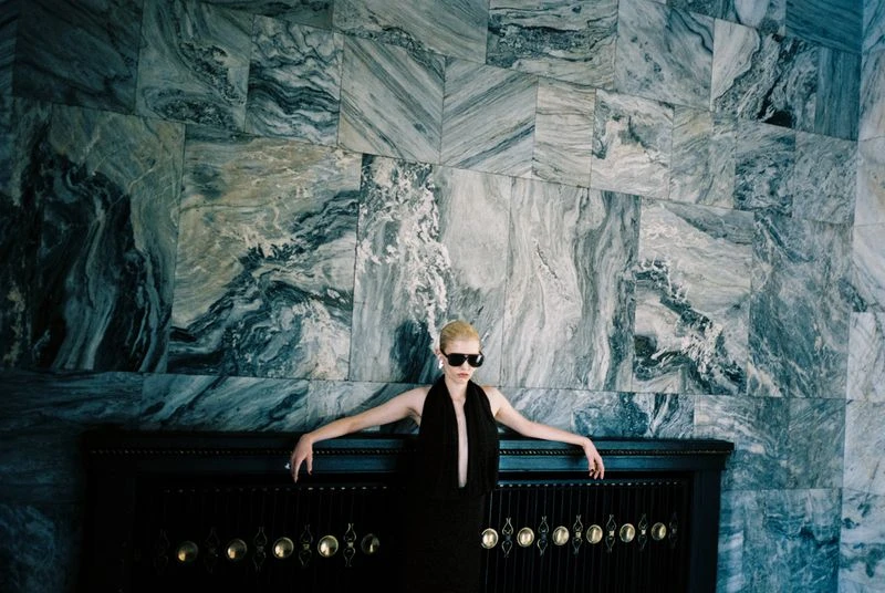 Fashion shot of an adult female with blond hair and sunglasses, wearing a black dress, posed in an indoor setting with a marble wall.