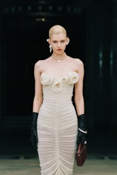 Model wearing a cream ruched bodycon gown with rosette bust detail, black leather opera gloves, and holding a burgundy clutch.