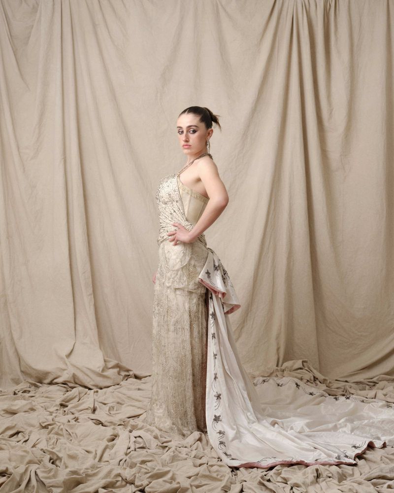 Woman in embellished gown poses in photo studio against draped backdrop, exhibiting fashion and elegance in formal attire.