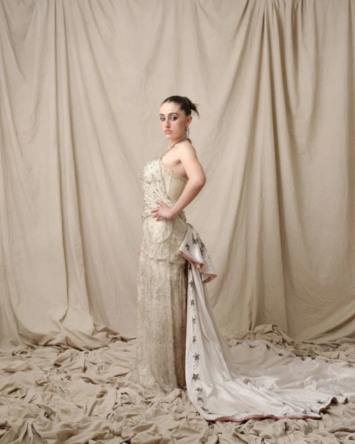 Woman in embellished gown poses in photo studio against draped backdrop, exhibiting fashion and elegance in formal attire.