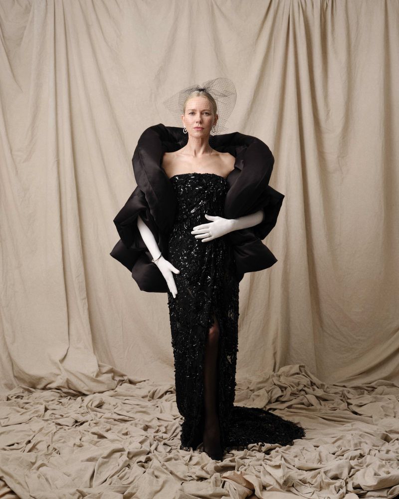 A woman in a black gown with large shoulder accents poses against a draped backdrop. She wears white gloves and a black hairpiece.