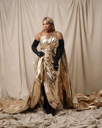 Serena Williams poses in a gold gown against a beige backdrop, wearing long black gloves and a golden headpiece in a studio setting.