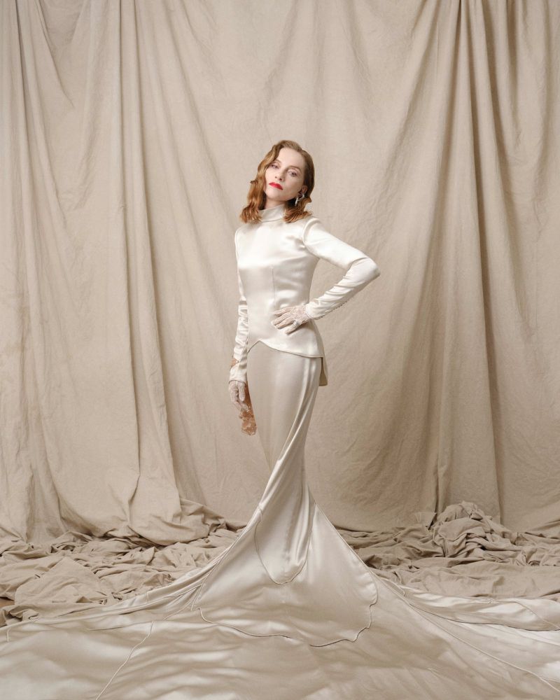 Woman in long-sleeved white gown standing against a draped fabric background in a fashion photoshoot, looking directly at the camera.