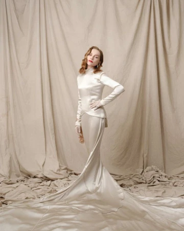 Woman in long-sleeved white gown standing against a draped fabric background in a fashion photoshoot, looking directly at the camera.