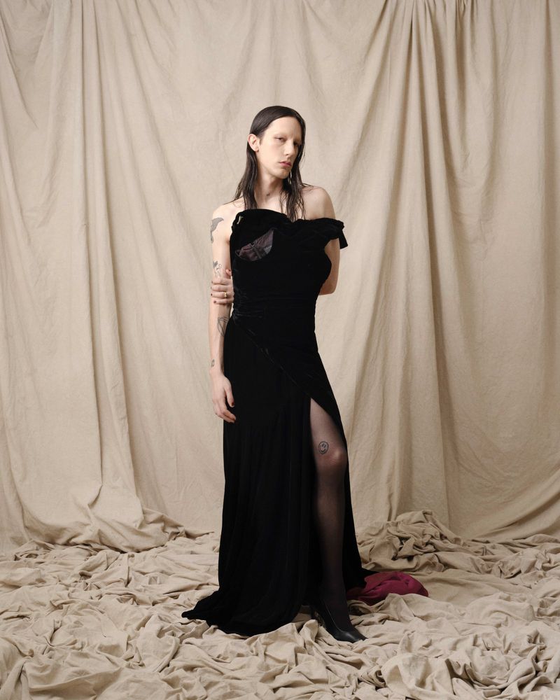 Elegant adult female in a black velvet dress with a thigh-high slit poses in front of a neutral drape in a studio setting.