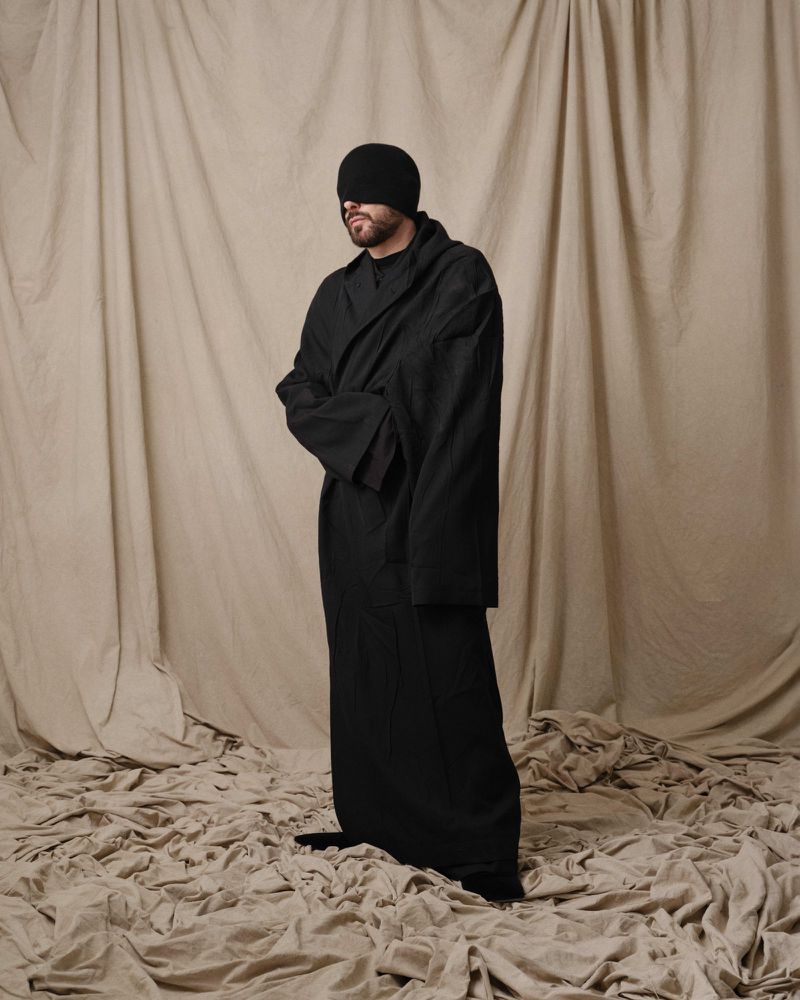 Full-length studio shot of a bearded adult male wearing a black robe and beanie, standing against a beige fabric background and floor.