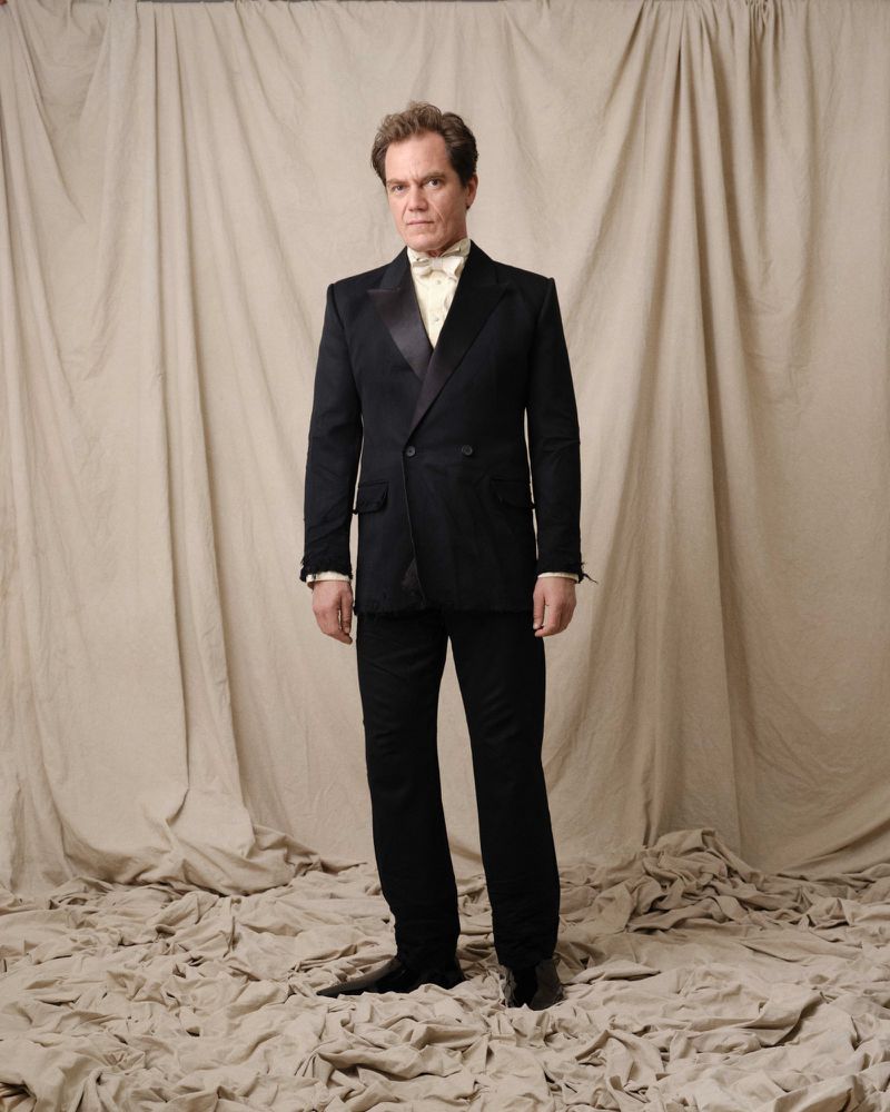 Full-length portrait of an adult male in a black tuxedo with a white bow tie, standing against a draped beige backdrop, formal attire.