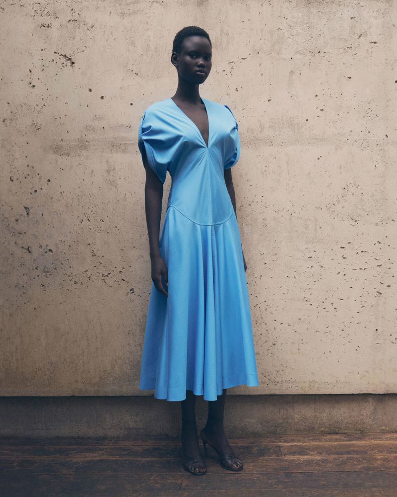 Model Nyakong Chan wearing a sky blue midi dress with a plunge neckline standing against a concrete wall.
