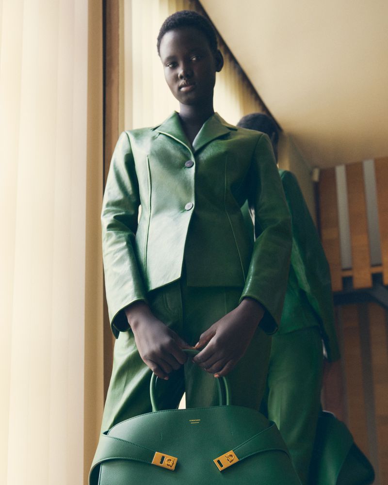 Fashion shot of an African woman in a green suit and matching handbag, standing indoors with a reflective surface visible.