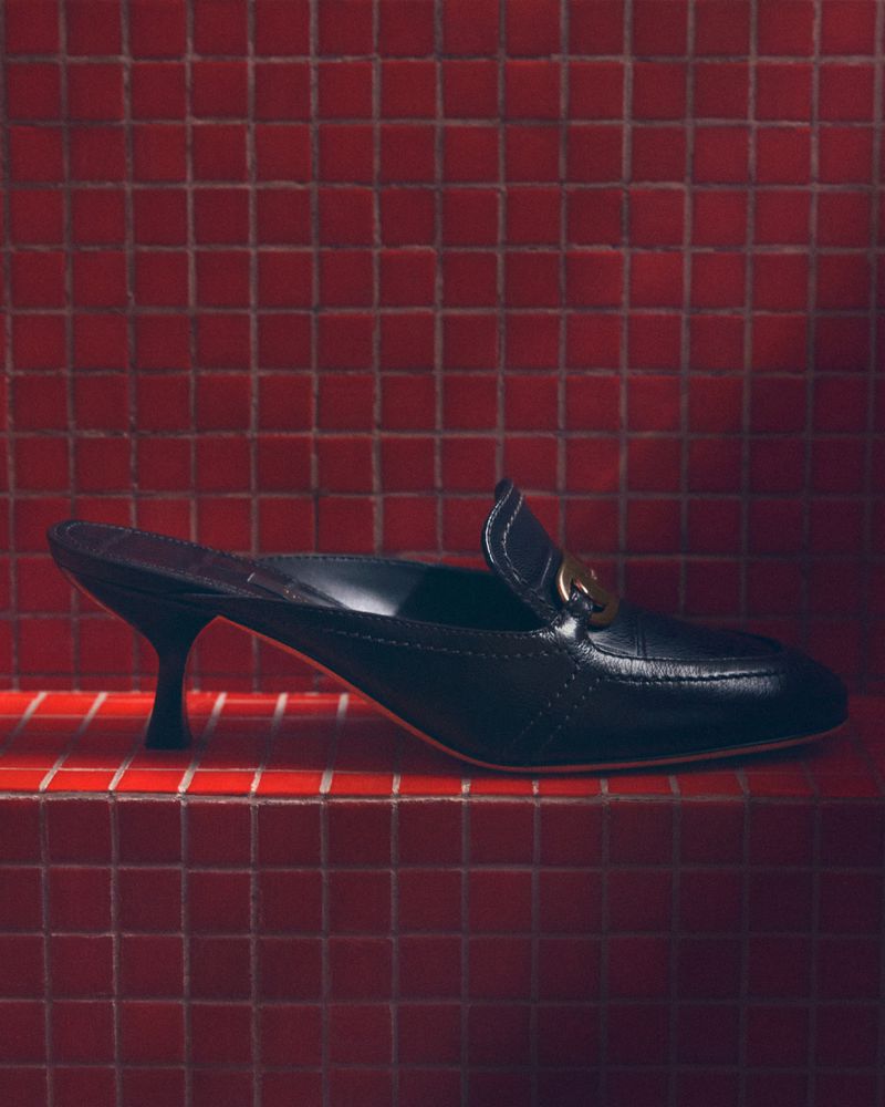 Black leather mule heel with a gold logo detail on a red tiled surface creating a visually striking fashion product shot.