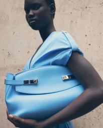 Model holding a light blue handbag wearing a matching blue dress against a textured concrete wall background